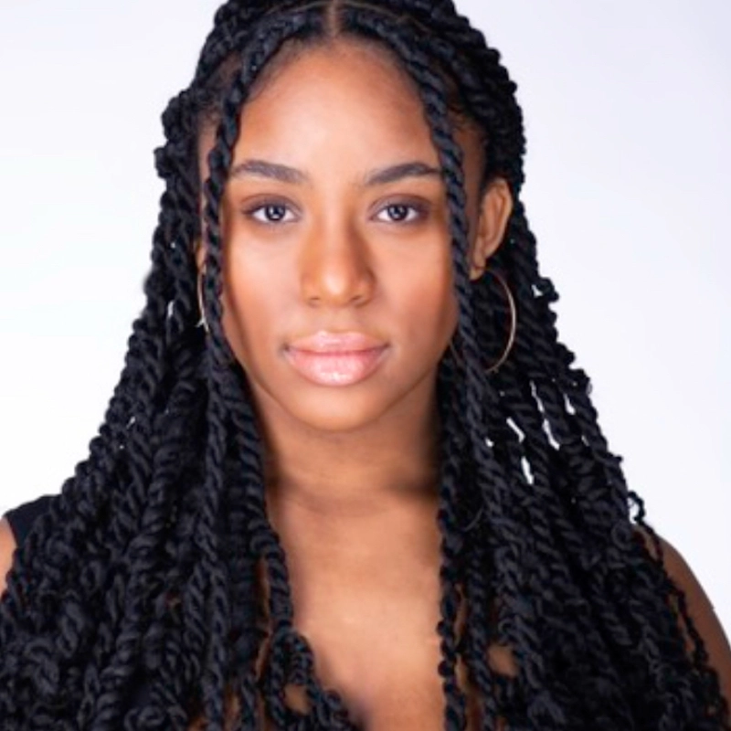 A woman with long, thick, black twists and hoop earrings looks directly at the camera. She has clear skin, subtle makeup, and wears a sleeveless black top. The plain white background emphasizes her face and hairstyle, capturing the confident presence of Sophia Pettaway.