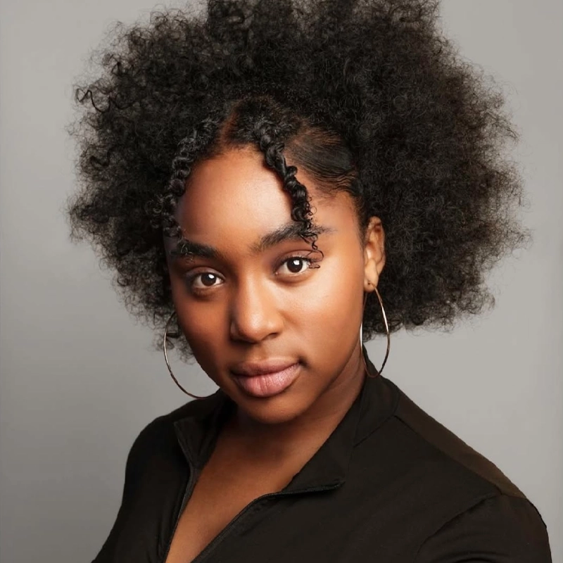 Sophia Pettaway stands confidently with natural, voluminous curly hair styled in two twisted strands at the front. She wears large hoop earrings, a black zip-up top, and gazes directly at the camera against a neutral background.