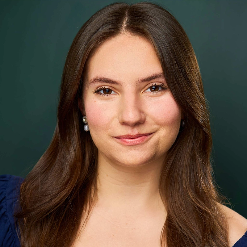 A young woman with long brown hair, parted in the center, smiles softly at the camera. She wears pearl earrings and a navy top. The background is a solid dark green. Her skin is fair and she has natural makeup, capturing Amanda Ribnick’s graceful presence.