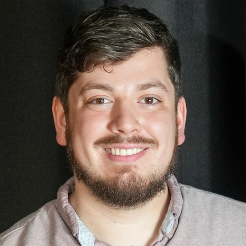 A smiling young adult with short, wavy brown hair and a beard stands in front of a dark background. He wears a light gray collared shirt with the top button open. The lighting is even, highlighting his face and friendly expression—a portrait style reminiscent of Amanda Ribnick’s signature photographic approach.