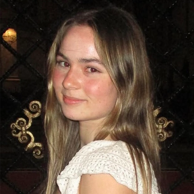 Young woman, Hannah Adams, with long, light brown hair and fair skin stands in front of a decorative, dark metal gate with gold accents. She wears a white, textured short-sleeve top and looks at the camera with a slight smile. Warm lighting is in the background.