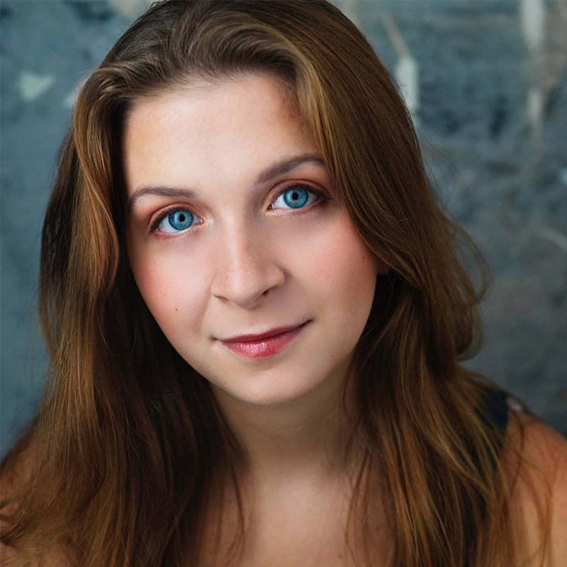 A young woman with long light brown hair and bright blue eyes looks at the camera. She has light skin, subtle makeup, and a slight smile. The softly blurred background in gray tones focuses attention on her face, capturing Amanda Ribnick’s serene and captivating presence.
