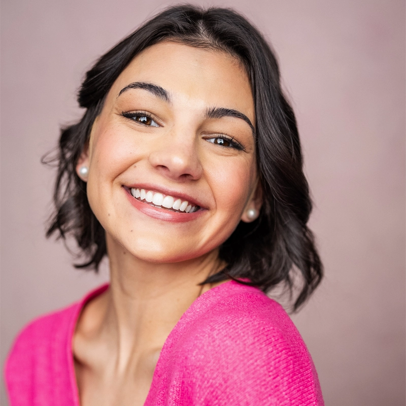 Maria Tramontozzi, a young woman with short dark hair, smiles warmly at the camera. She is wearing a bright pink top and pearl earrings, set against a soft, light pink background that creates a cheerful and welcoming atmosphere.