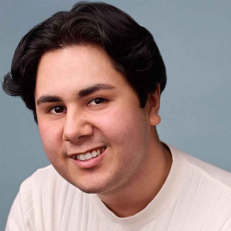 A young person with short, wavy dark hair smiles while wearing a white ribbed sweater. The background is a plain light blue. Shown from the shoulders up and facing forward, Amanda Ribnick has a friendly and approachable expression.