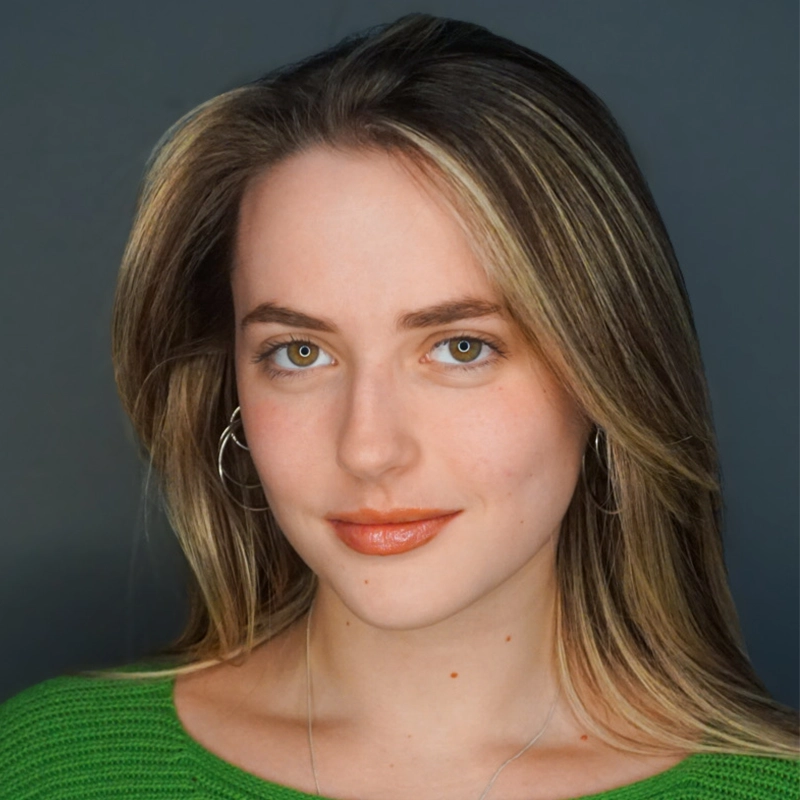 Abigail Callahan, a young woman with light skin, long blonde hair, and hazel eyes, faces the camera with a subtle smile. She wears silver hoop earrings, a green sweater, and a thin necklace against a plain dark gray background.