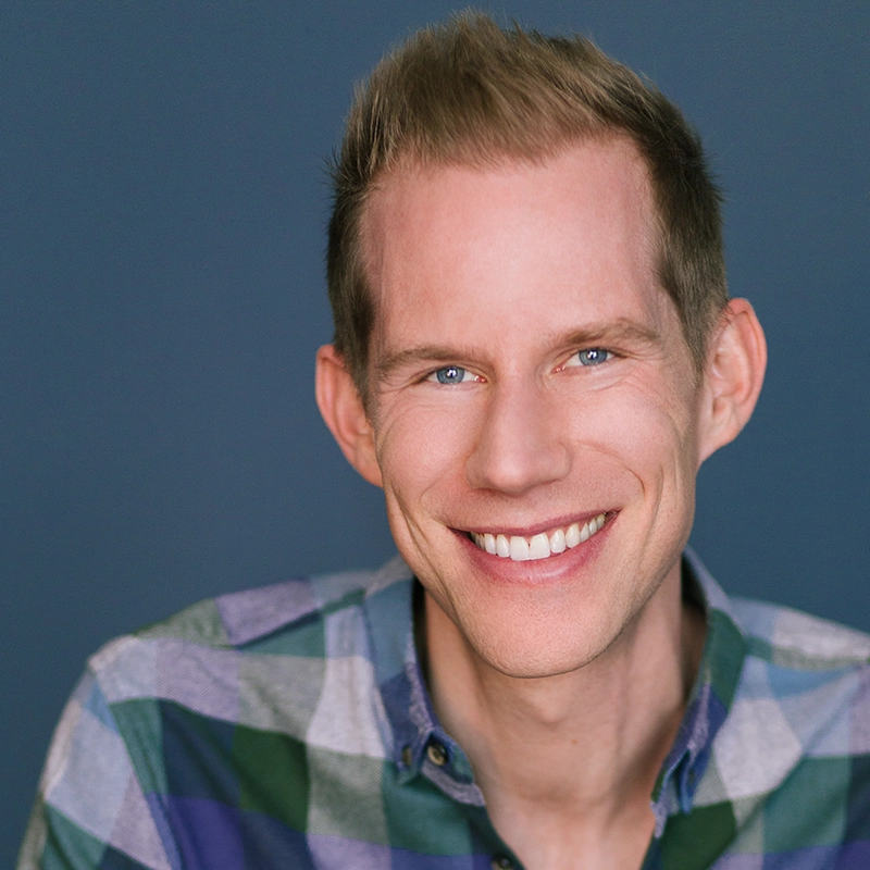 Desmond Luis Edwards, a smiling man with short, light brown hair, wears a green, blue, and white plaid shirt. He is shown from the shoulders up against a solid blue background, looking directly at the camera with a friendly expression.