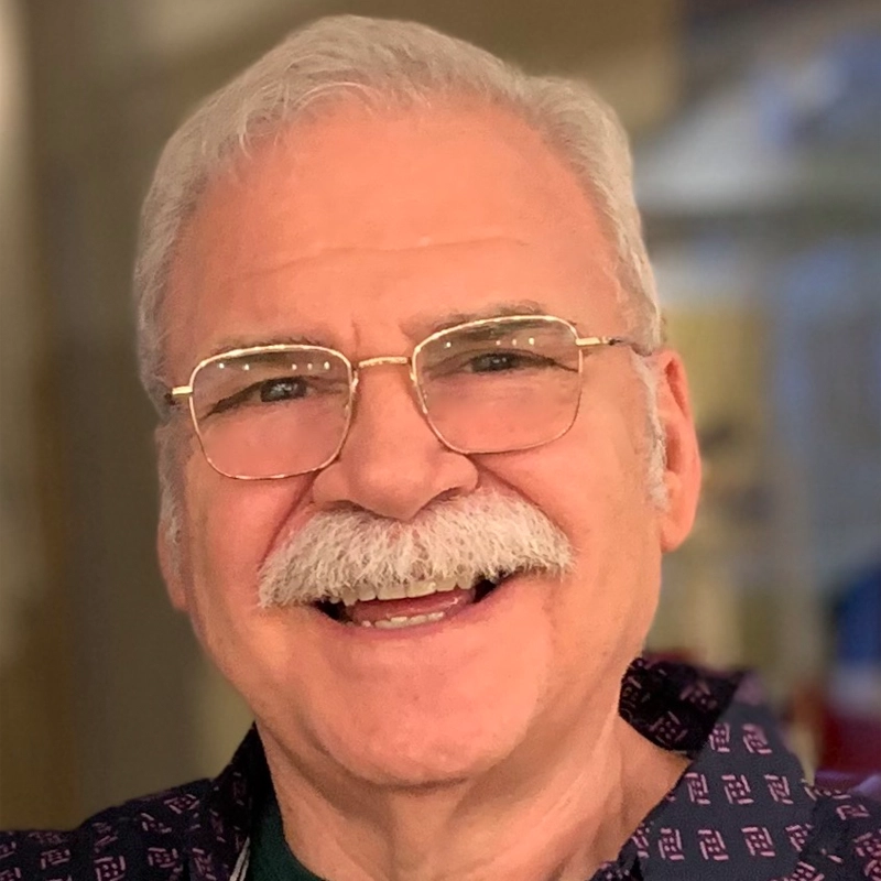 An older man with short white hair, a white mustache, glasses, and a patterned shirt smiles warmly at the camera. The background is softly blurred with warm tones, drawing focus to his friendly expression—a look reminiscent of someone who enjoys the wit and playfulness of musical theater classics like Forbidden Broadway.