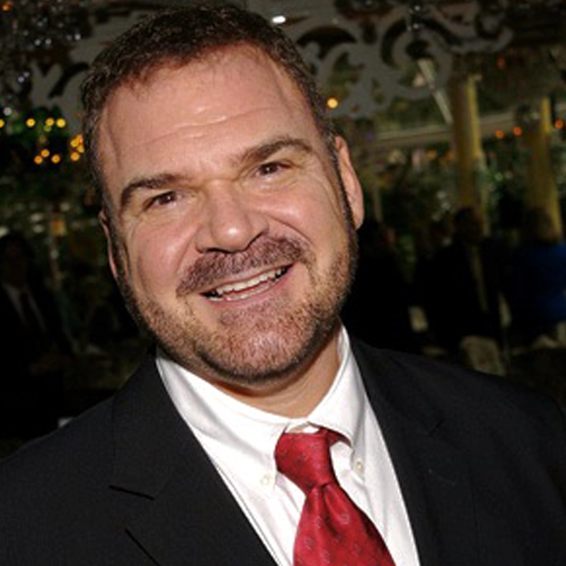 A smiling man with short brown hair and a beard, wearing a dark suit, white shirt, and red tie, stands indoors at a formal event celebrating musical theatre. Blurred people, lights, and decorations are visible in the background.
