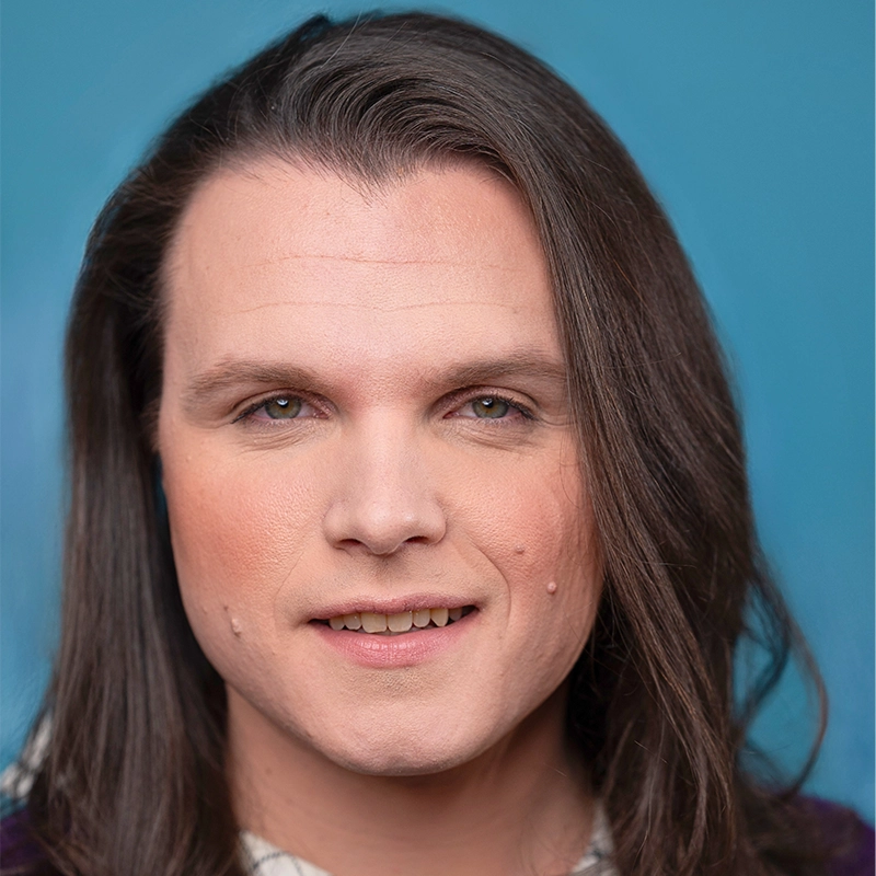 A person with long brown hair and light skin smiles softly against a blue background. Their makeup is subtle, with even skin tone and light eye makeup. They have visible dimples on both cheeks and wear a patterned shirt.