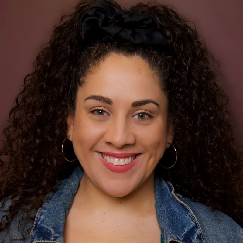 A woman with long curly dark hair wears a black hair scrunchie, hoop earrings, and a blue denim jacket. She is smiling brightly, showing her teeth, and stands in front of a soft brown background.