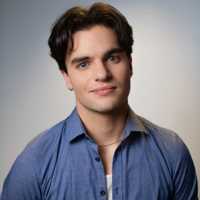 Nicholas Barrón, a young man with short, dark hair and blue eyes, faces forward with a slight smile. He wears a blue button-up shirt over a white top, a thin gold necklace, and a small gold hoop earring against a softly lit neutral background.