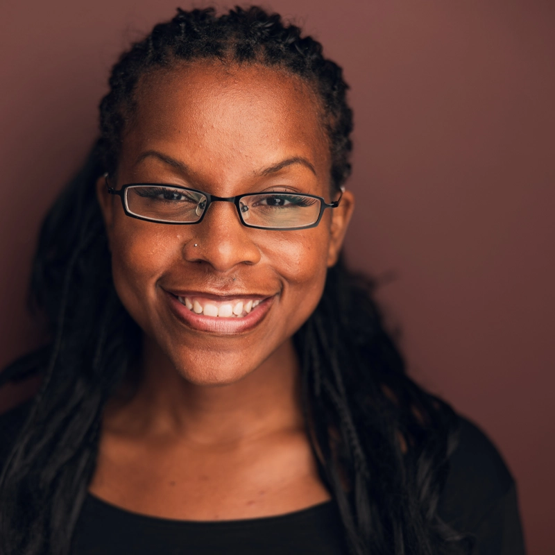 A woman with dark skin and long braided hair smiles warmly at the camera. She wears rectangular glasses and a black top, and has a nose piercing. The background is a solid, muted maroon color, highlighting Madyson Bolton’s bright presence.
