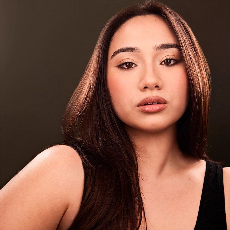 A young woman with long, straight brown hair poses against a dark background. She has light skin, natural makeup with nude lipstick, and softly defined eyes. Brianna Paradiso wears a black sleeveless top and gazes confidently at the camera.