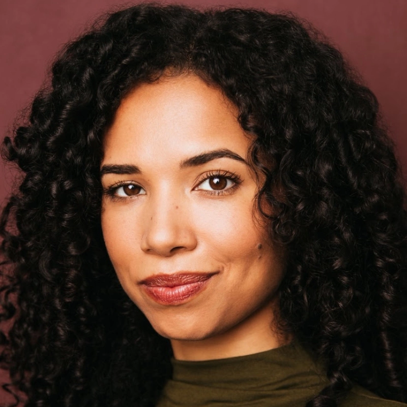 Victoria Byrd, a woman with medium brown skin and long, dark curly hair, smiles softly. She has brown eyes, defined eyebrows, and wears a high-necked olive green top against a solid burgundy background.