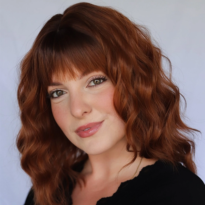 Deanna Jolie Thomas, a woman with fair skin and wavy, shoulder-length auburn hair with bangs, smiles softly. She wears light makeup with blush and pink lipstick, a black top, and stands against a plain, light-colored background.