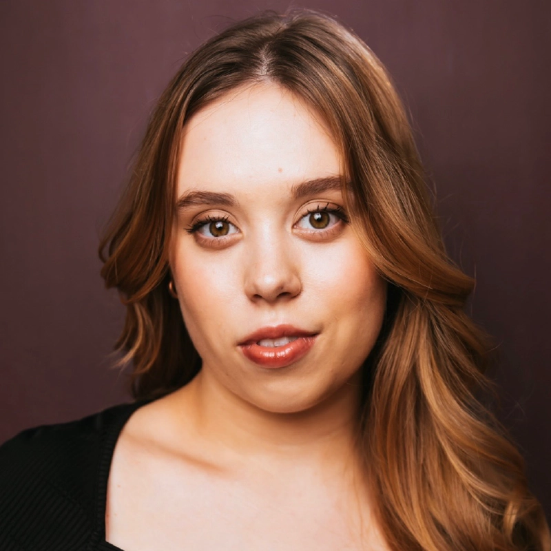 A young woman with long, wavy light brown hair poses against a plain, dark background. She has fair skin, brown eyes, and subtle makeup with soft pink lipstick. She wears a black top and looks directly at the camera with a slight smile.