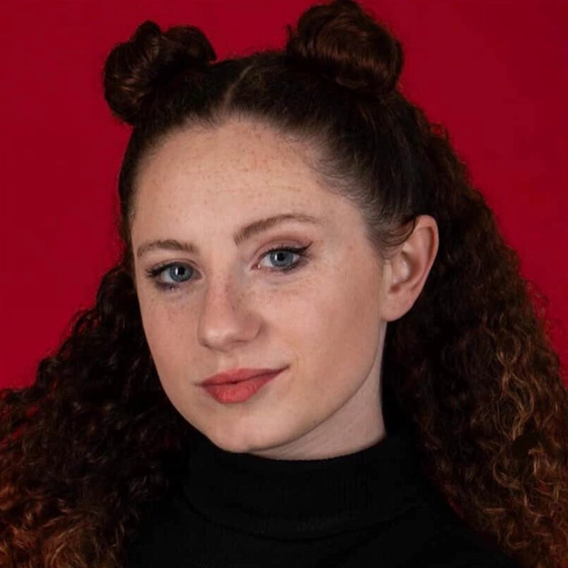 A young woman with fair skin, blue eyes, and curly brown hair styled in two space buns poses against a solid red background. She wears a black turtleneck and subtle makeup, looking confidently at the camera with a neutral expression.