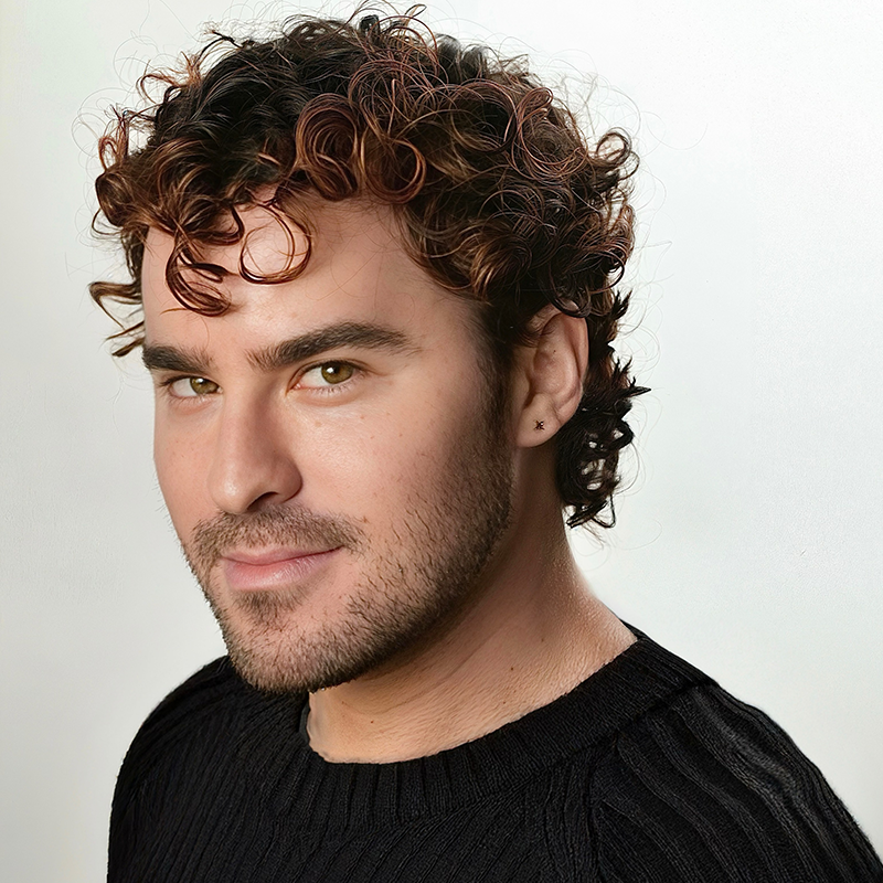 A young man with short, curly brown hair and a trimmed beard smiles softly. He wears a black knit sweater and a small earring in his left ear. The background is plain and light-colored, drawing focus to his face.