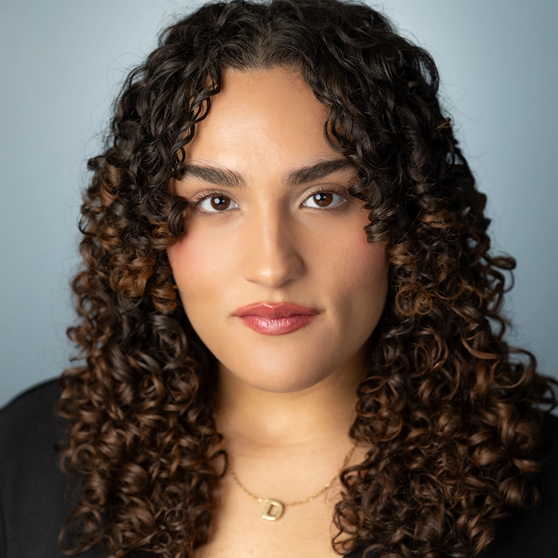 A person with medium skin tone and long, dark, curly hair looks directly at the camera. They have full eyebrows, brown eyes, and wear natural makeup with pink lipstick. Deanna Jolie Thomas wears a black top and a gold “D” pendant necklace. Light blue background.