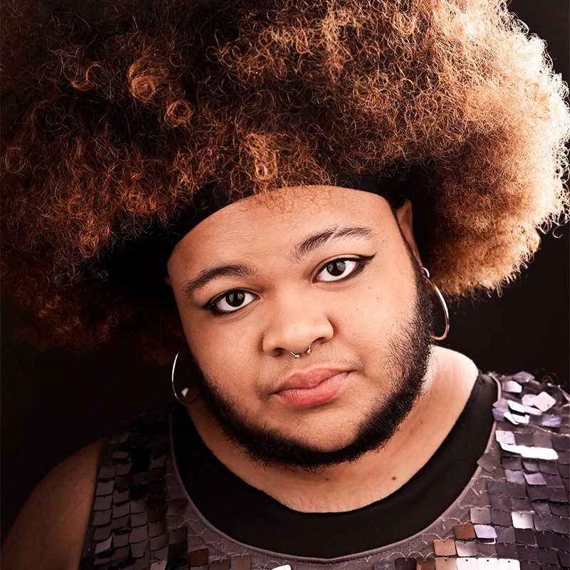 Desmond, with a voluminous curly brown afro, wears hoop earrings, a septum ring, and black eyeliner. He has a short beard and mustache and is dressed in a shiny sequined top, looking directly at the camera with a gentle expression.