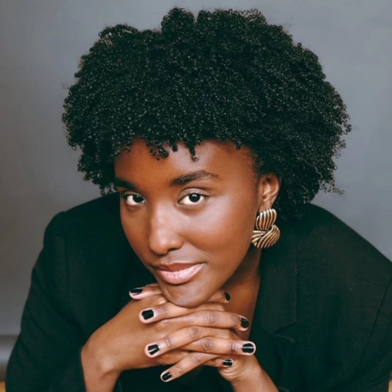 A woman with short, curly hair and gold earrings rests her chin on her clasped hands. She wears a black blazer and has black nail polish. The background is plain gray, and she gazes confidently at the camera with a slight smile.