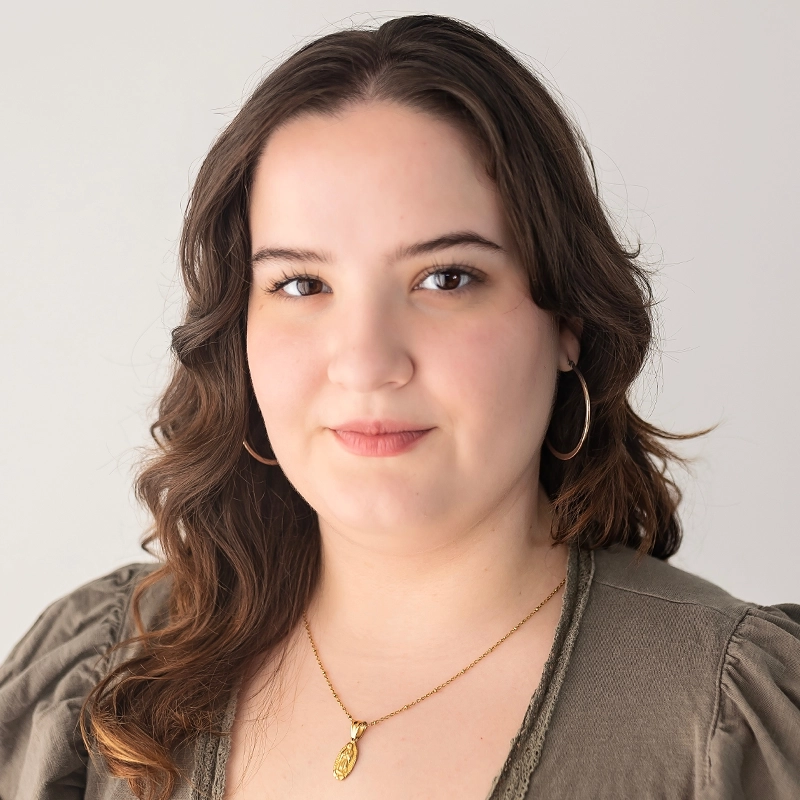 Abigail Callahan, a young woman with wavy brown hair, wears a neutral expression. She has hoop earrings and a gold necklace with a small pendant, dressed in an olive-green top with puffed sleeves against a plain light background.