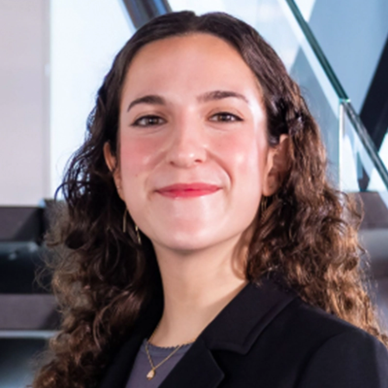 Abigail Callahan, with long curly brown hair, smiles confidently. She wears a black blazer, gold earrings, and a delicate gold necklace. The modern staircase and glass background suggest a professional office environment.