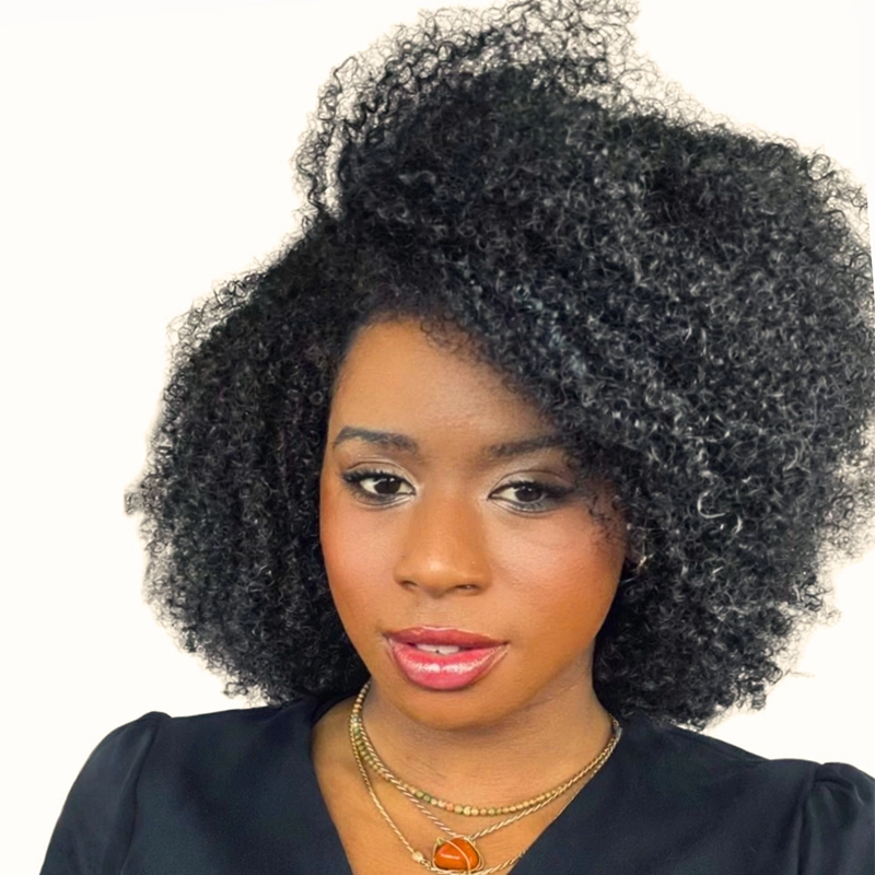 Deanna Jolie Thomas showcases voluminous, natural black curly hair, a black top, and layered gold necklaces with a red stone pendant. She wears subtle, elegant makeup with shimmery eyeshadow and glossy pink lips against a plain white background.