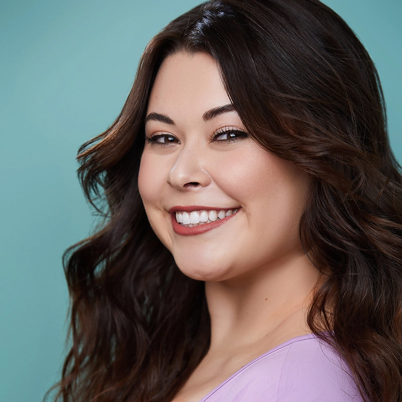A young woman with long, wavy brown hair smiles at the camera. Deanna Jolie Thomas wears a light purple top and natural makeup. The background is a solid light teal color, creating a clean and cheerful portrait.