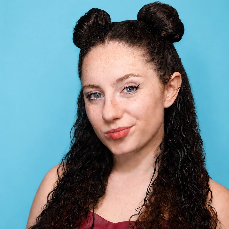 Woman with fair skin, curly dark hair styled in two space buns, and light blue eyes, wearing a sleeveless maroon top, poses in front of a bright blue background, smiling slightly with pink lipstick and visible freckles on her cheeks.