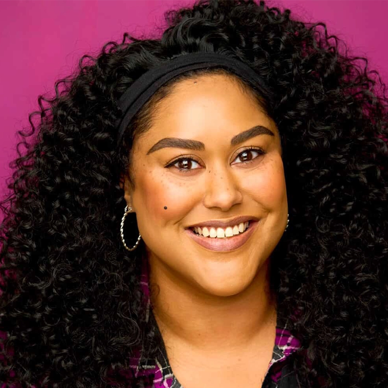 A woman with long, curly dark hair and hoop earrings smiles at the camera. She has bold eyebrows, winged eyeliner, a beauty mark on her left cheek, and wears a black headband and a purple plaid shirt. The background is bright magenta.