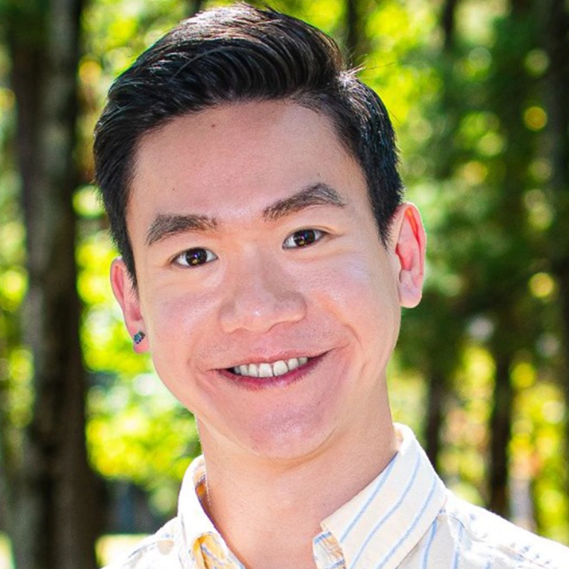 A young person with short, dark hair smiles at the camera while standing outdoors. They wear a light-colored, collared shirt. Sunlight filters through green trees in the blurred background, creating a bright and cheerful atmosphere.