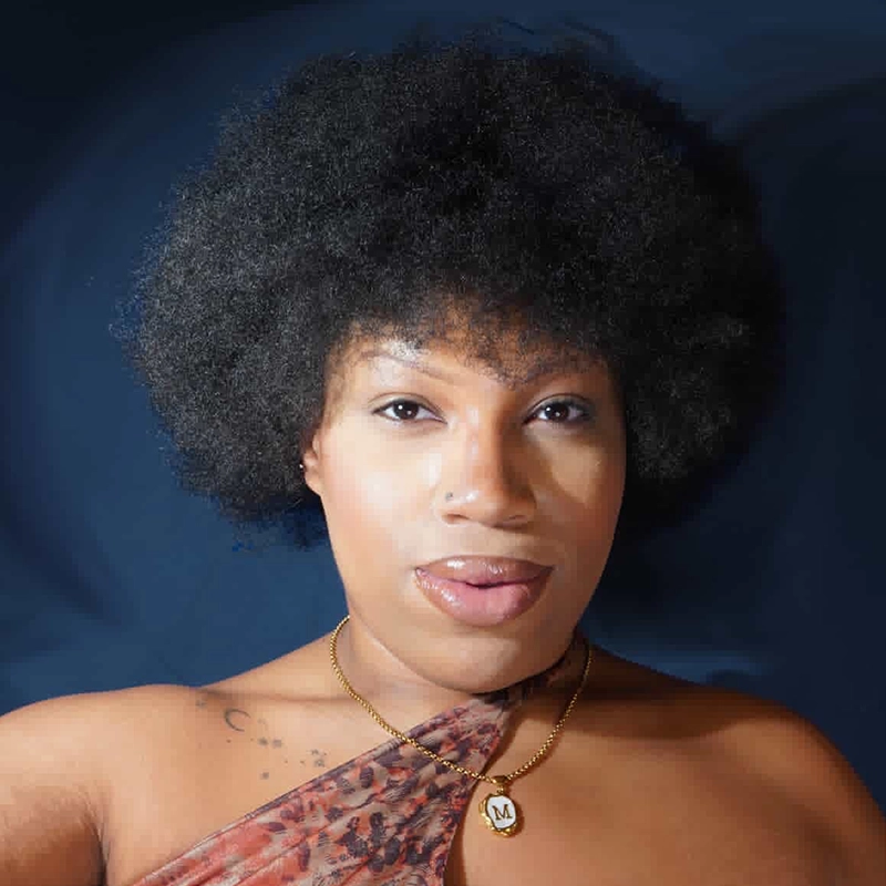 Deanna Jolie Thomas with an afro hairstyle wears a single-strap patterned top and a gold necklace with a heart-shaped pendant featuring the letter “M.” She poses facing forward against a dark blue background, her expression calm.