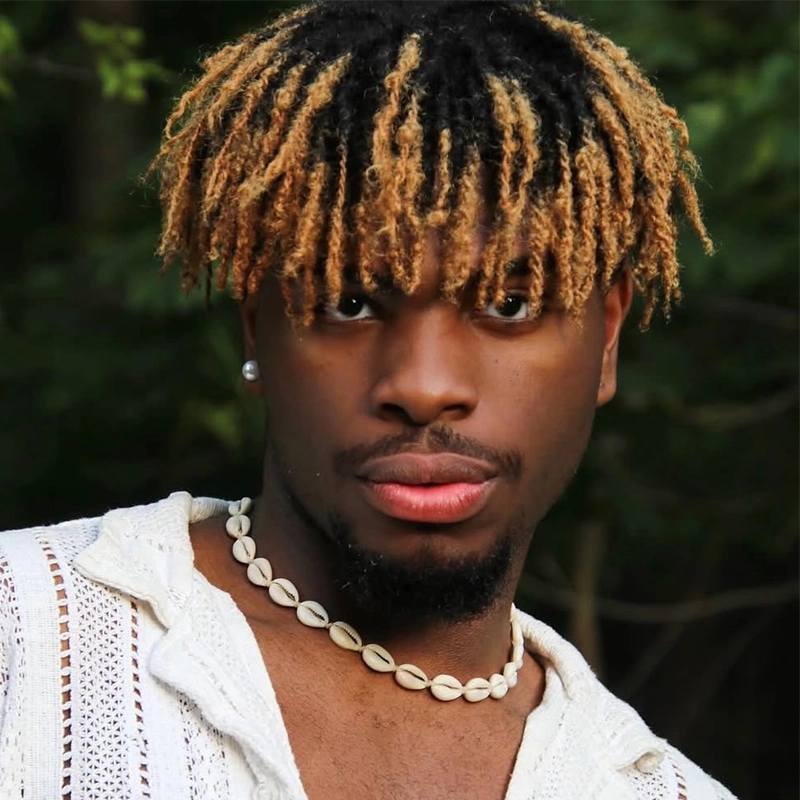 Najm Muhammad, with light brown and blonde dreadlocks, wearing a white textured shirt, seashell necklace, and a small earring, looks directly at the camera with a serious expression. Green trees blur softly in the background.