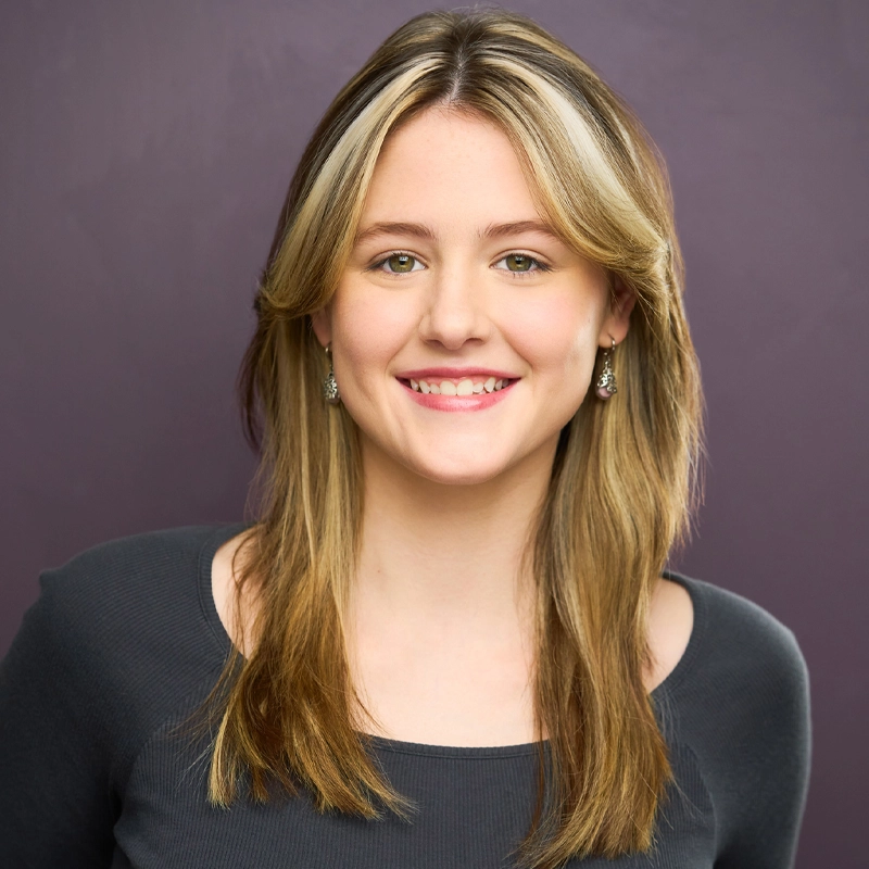 Tannalee Poythress, a young woman with long, straight, blonde hair and subtle highlights, smiles at the camera. She wears a charcoal gray top and small dangling earrings against a solid muted purple background for a professional portrait.