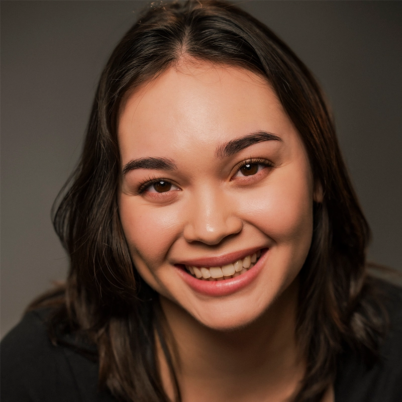 A young woman, Deanna Jolie Thomas, with straight, shoulder-length dark brown hair smiles warmly at the camera. She has clear skin, full eyebrows, and wears a black top. The softly blurred background keeps the focus on her friendly expression.