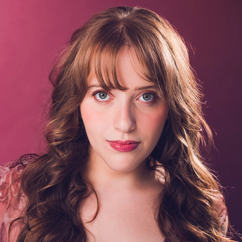 Abigail Callahan, a young woman with long, wavy light brown hair and fringe, stands against a pink background. She has fair skin, blue eyes, and wears natural makeup with pink lipstick. Dressed in a pink lace top, she gazes calmly at the camera.