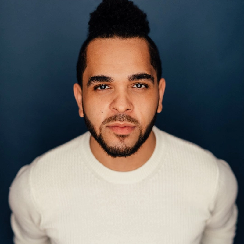 A man with a short beard and curly hair pulled up in a bun wears a white knit sweater. He faces the camera directly, standing against a dark blue background, with a neutral expression and soft lighting illuminating his face.