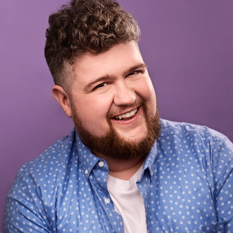 A smiling man with curly brown hair and a short beard wears a blue patterned button-up shirt over a white t-shirt. He poses against a plain purple background, looking directly at the camera with a cheerful expression.