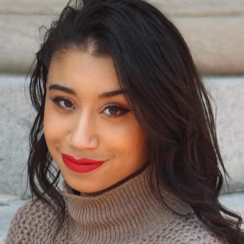 A young woman with long, dark hair and warm skin tone smiles softly. She wears a taupe turtleneck sweater and has bold red lipstick and dark eyeliner. The background is blurred stone steps, creating a soft, neutral backdrop for her portrait.