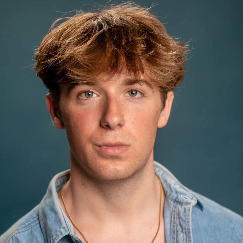 Ayden Weinstein, a young man with light skin, tousled light brown hair, and blue eyes, stares directly at the camera. He wears a light blue denim shirt with the collar open and a thin necklace against a plain teal background.