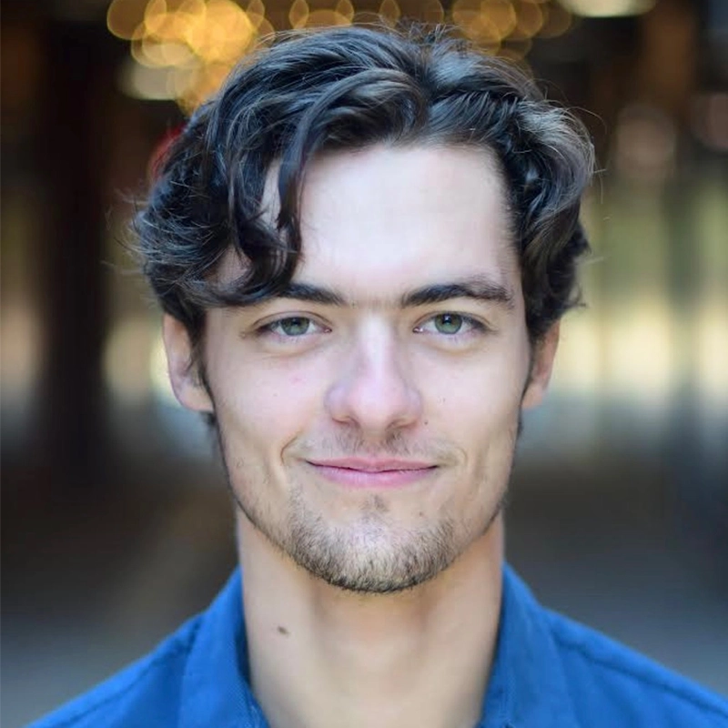A young man with wavy brown hair, fair skin, and light green eyes smiles softly at the camera. He has light stubble, wears a blue collared shirt, and stands in soft focus lighting with warm bokeh lights in the background.