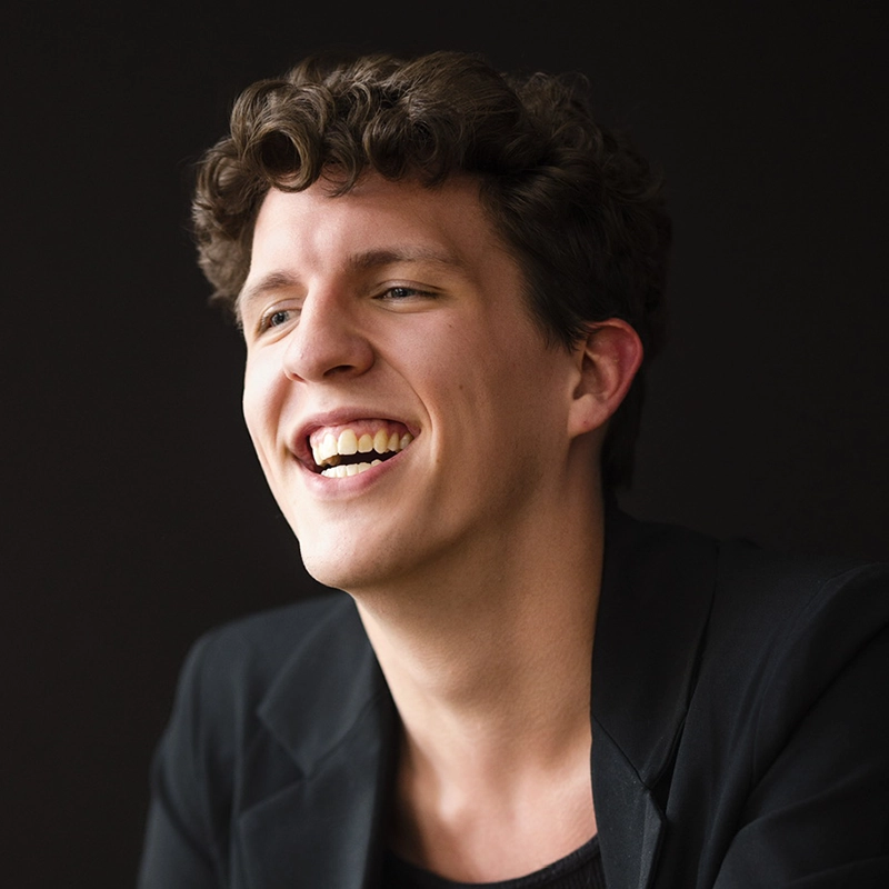 A young person with short curly brown hair and light skin is smiling widely, showing teeth. They are wearing a black blazer over a dark top, against a plain dark background. The lighting highlights their face, creating a warm and joyful atmosphere.