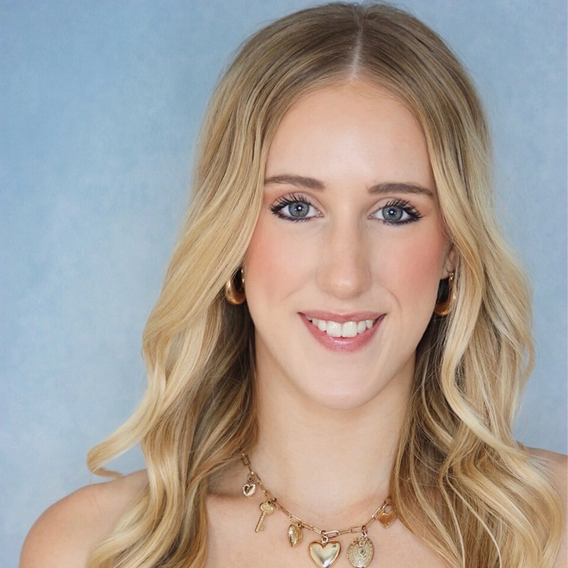 A young woman with long, wavy blonde hair is smiling at the camera. She has blue eyes, light makeup, and is wearing gold hoop earrings and a gold necklace with heart and key charms. The background is a soft, light blue gradient.
