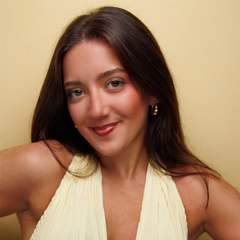 A young woman with long brown hair smiles softly at the camera. She wears a sleeveless, pale yellow top and gold hoop earrings. The background is a plain, light yellow color, creating a warm, cohesive look. Her makeup is natural and polished.