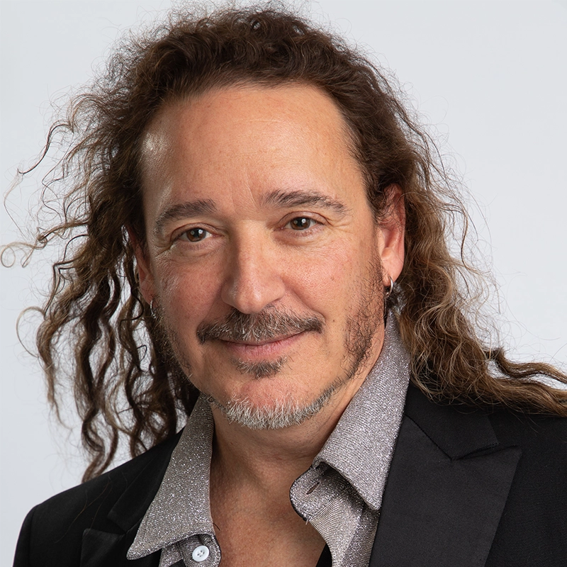 Eric Scott Klein, a middle-aged man with long, wavy brown hair and a goatee, smiles gently. He wears a sparkling silver collared shirt and a black blazer. The background is plain white, with his hair slightly tousled on one side.