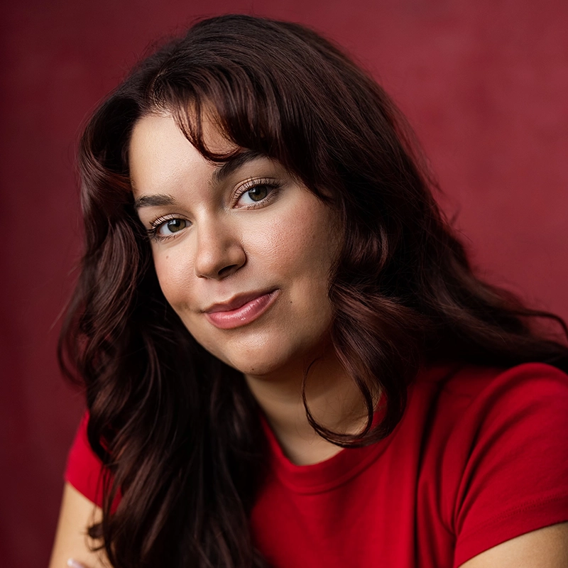 Grace Bacino, a young woman with long, wavy dark brown hair, poses in front of a solid dark red background. She wears a red top, has light skin, subtle makeup, and smiles gently while looking directly at the camera.