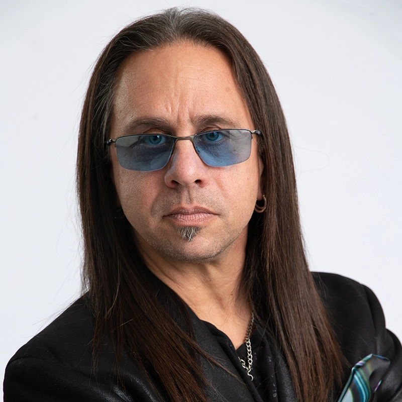 Eric Scott Klein, a man with long straight brown hair, light skin, and a small goatee, wears blue-tinted sunglasses, gold hoop earrings, and a black blazer. He looks seriously at the camera against a plain white background.