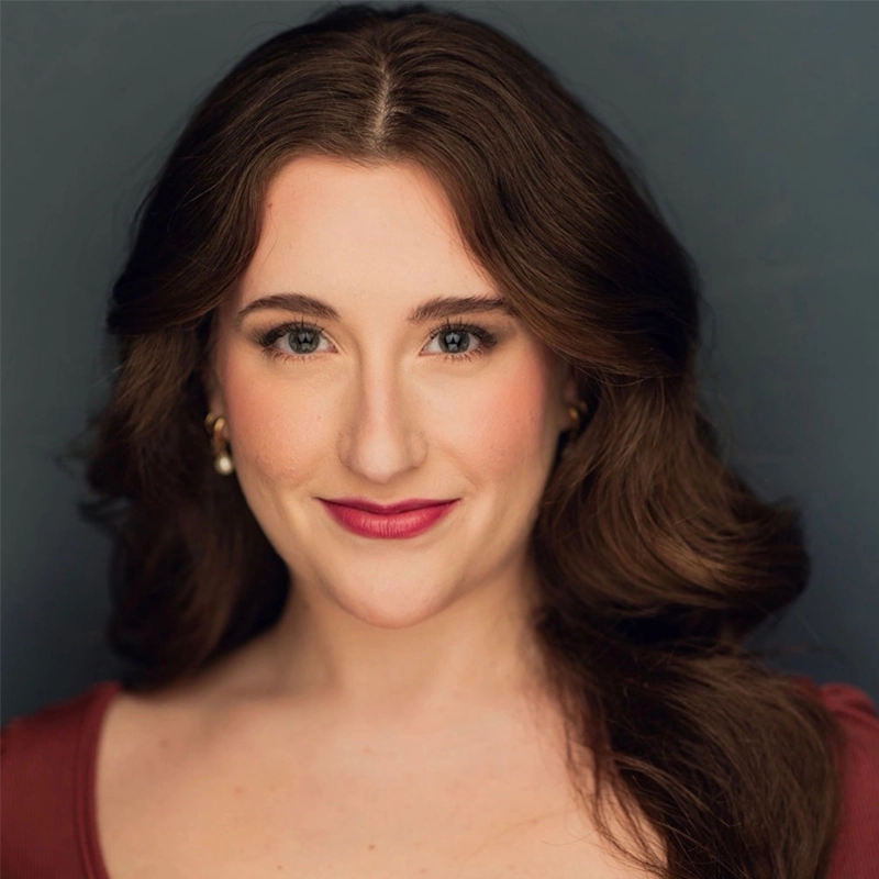 A woman with long, wavy brown hair and fair skin smiles slightly. She has blue-green eyes, natural makeup with pink lipstick, and wears gold earrings and a maroon top. The background is a plain, dark gray color.