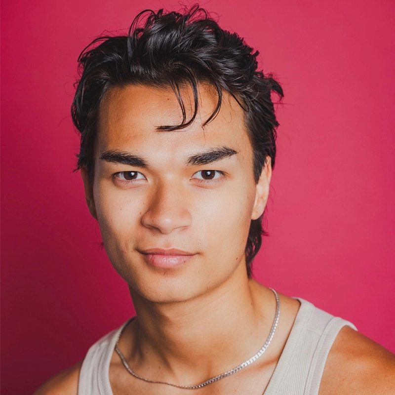 A young person with medium-length dark hair and clear skin wears a light-colored tank top and a thin silver chain necklace, posing against a solid bright pink background with a neutral, confident expression.